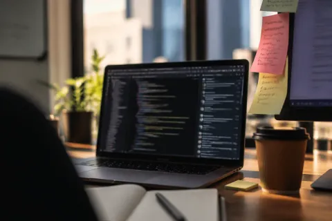 Foto editorial em close-up da mesa de um desenvolvedor, tela do notebook com um editor de código desfocado ao lado de uma janela de chat, post-its coloridos na borda do monitor com nomes de comandos escritos à mão, luz de fim de tarde
