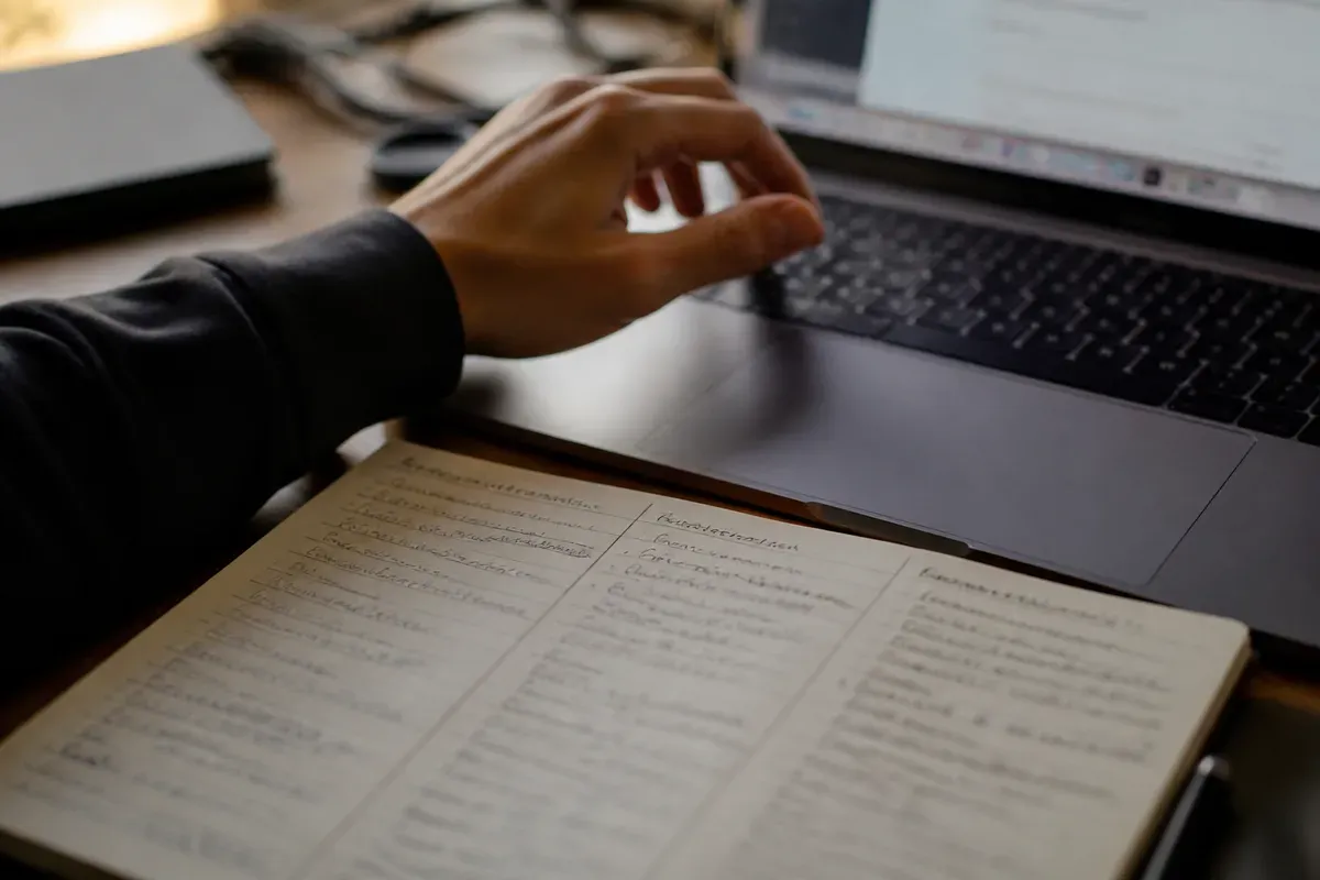 Close-up de uma mão sobre o teclado de um notebook ao lado de um bloco de notas com três colunas de anotações, luz suave de fim de tarde